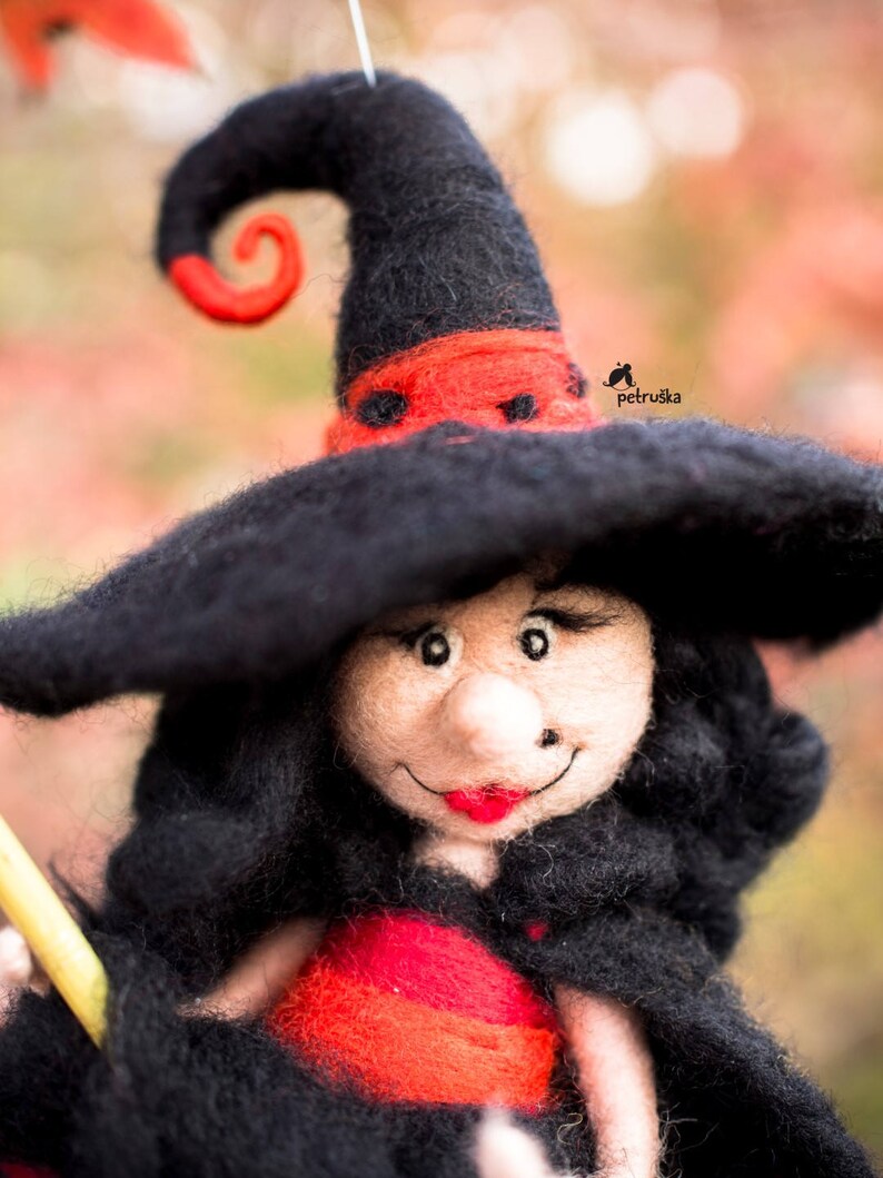May include: A felt witch doll with a black hat and red trim, holding a broom. The doll has a smiling face with red lips and black eyes.