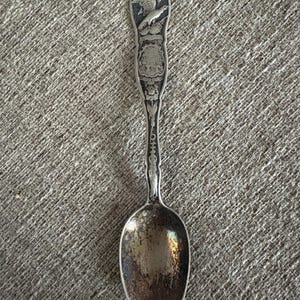 May include: A vintage silver spoon with an ornate handle featuring an eagle and shield design. The spoon's bowl shows signs of wear, with a tarnished appearance. The handle has detailed floral and scrollwork.