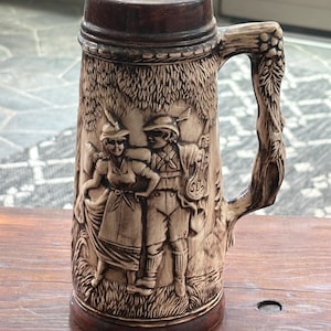 May include: A tall ceramic beer stein featuring a relief design of a person in a dress and a person in shorts and a hat standing in a forest. The handle is textured to resemble a tree branch with leaves. The stein has a dark brown rim and base.