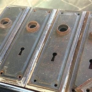 May include: A set of four rectangular metal door plates with a dark, weathered finish. Each plate features a central circular knob opening, a keyhole cutout below, and small screw holes at the top and bottom corners, showing signs of oxidation.