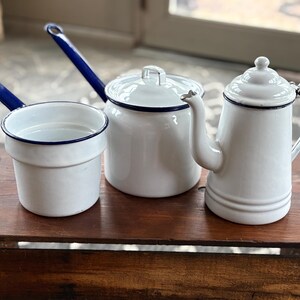 Op de afbeelding: Een verzameling witte emaille keukenartikelen, waaronder een kleine steelpan met blauwe handgreep, een pot met deksel en een theepot. Elk stuk heeft een blauwe rand en wordt gepresenteerd op een houten oppervlak. De set heeft een vintage uitstraling.