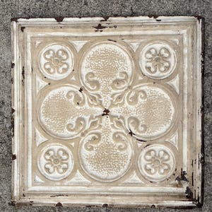 May include: Square, off-white decorative metal tile with a repeating floral and circular pattern. The tile has a distressed, aged appearance with visible rust and wear on the edges. The design features raised, textured details.