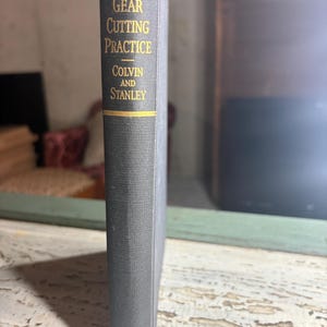Könnte beinhalten: Ein dunkelgraues Hardcover-Buch mit dem Titel Gear Cutting Practice von Colvin und Stanley, herausgegeben von der McGraw-Hill Book Company. Der Buchrücken zeigt goldene Schriftzüge und horizontale goldene Linien mit dem Titel.