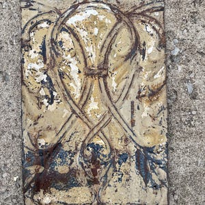May include: A rectangular, antique metal decorative panel with a faded, distressed finish. The panel features a symmetrical design of intertwined floral or leaf-like shapes in cream, brown, and blue hues. The metal shows signs of rust and paint chipping.