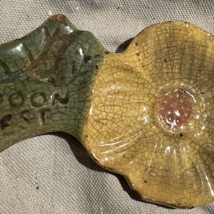 May include: A ceramic spoon rest in the shape of a leaf and flower. The leaf section is green with the words "Spoon Rest" in black. The flower section is yellow with a pink center and a cracked glaze.