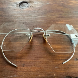 Può includere: Occhiali vintage senza montatura con lenti ottagonali e montatura in metallo argentato. Gli occhiali hanno aste curve e piccoli naselli colorati. Le lenti sono trasparenti, su uno sfondo di legno.