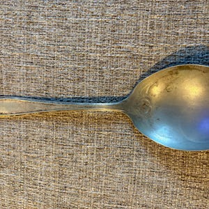 Puede incluir: Una cuchara de servir plateada con un cuenco ovalado y un mango largo. El mango tiene un diseño decorativo cerca del extremo, y el cuenco tiene un patrón floral. La cuchara es probablemente vintage o antigua.