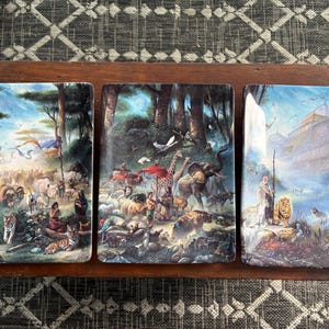 May include: A set of three decorative ceramic tiles depicting biblical scenes. The tiles are set in a wooden tray. The scenes include animals, people, and landscapes, with vibrant colors and detailed artwork.