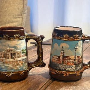 May include: Two ceramic beer steins featuring dark brown rims and bases with ornate gold-toned filigree detailing. The left stein depicts a white building by water, while the right stein shows a historic building with a tall column in a city square.