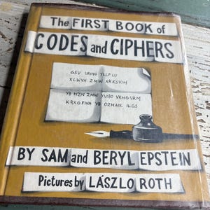 May include: A hardcover book titled The First Book of Codes and Ciphers by Sam and Beryl Epstein with pictures by László Roth. The mustard yellow cover features a white paper graphic with encoded text, a black inkwell, and a white quill pen.