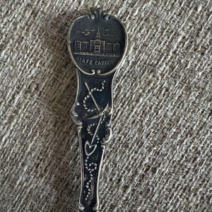 Pode incluir: Uma colher de prata vintage com um design detalhado. A tigela da colher apresenta uma imagem de um edifício com o texto "STATE CAPITOL" abaixo. O cabo tem um padrão ornamentado com uma âncora, uma pá e outros elementos decorativos.