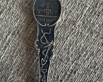 May include: A vintage silver spoon with a detailed design. The spoon's bowl features an image of a building with the text "STATE CAPITOL" below it. The handle has an ornate pattern with an anchor, a shovel, and other decorative elements.