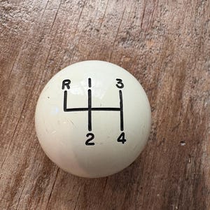 Puede incluir: Pomo de palanca de cambios color crema con letras negras. El pomo muestra el patrón de cambio de marchas: R, 1, 2, 3 y 4. El pomo redondo parece estar hecho de plástico duro o resina.
