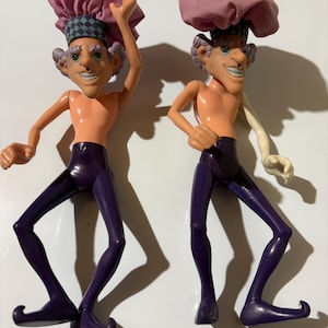 May include: Two vintage, whimsical figurines of a shirtless man with a smiling face, purple pants, and a pink hat. One figure has an arm raised, the other has an arm at its side. The figures have a unique, cartoonish design.