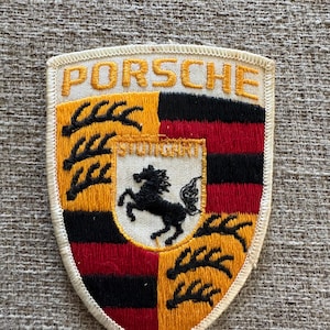 Könnte beinhalten: Gesticktes Porsche-Abzeichen mit dem Wort "PORSCHE" in goldener Schrift. Das Schilddesign hat schwarze, rote und goldene Abschnitte sowie ein schwarzes Pferdeemblem. Das Wort "STUTTGART" ist ebenfalls sichtbar.