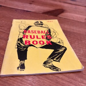 May include: A 1961 baseball rule book with a yellow cover featuring a black ink illustration of a baseball player in a crouched stance. The text BASEBALL RULES BOOK is printed in bold red capital letters across the center of the cover.