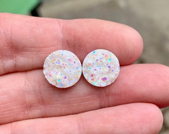 May include: Two round, white stud earrings with a sparkling, iridescent finish. The earrings have a textured surface with colorful flecks, creating a shimmering effect. These earrings are a simple yet elegant accessory.