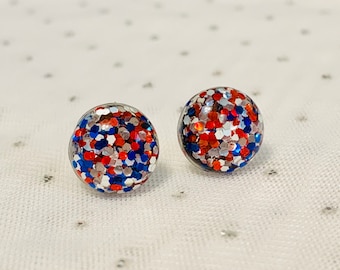 May include: A pair of stud earrings with red, white, and blue glitter embedded in clear resin.