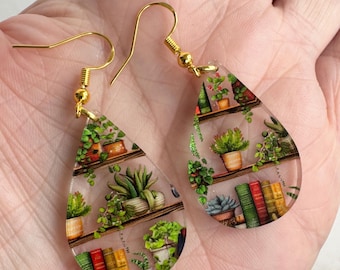 May include: A pair of teardrop-shaped earrings with gold hooks. The earrings are made of clear resin and feature a miniature bookshelf filled with books and potted plants.
