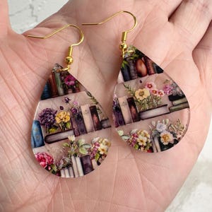 May include: A pair of teardrop-shaped earrings with a gold hook. The earrings are clear and feature a printed image of a bookshelf filled with books and flowers.