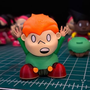 May include: A 3D-printed figurine of a cartoon character with orange hair, a green shirt, and red shoes. The figure has its arms raised and is sitting on a green grid surface. Other similar figurines are visible in the background.