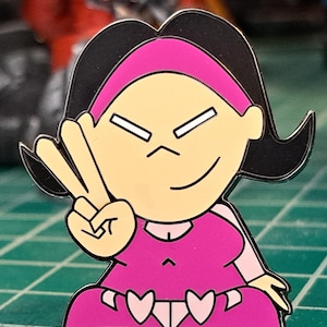 Peut inclure: Épinglette en émail colorée représentant un personnage de dessin animé avec des cheveux noirs, un bandeau rose et une robe rose. Le personnage fait le signe de la paix. L'épinglette a un contour argenté et trois cœurs en bas.
