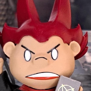 May include: A small figurine with a tan face, pointed red hair, and an angry expression featuring white eyes and a bared-teeth mouth. The character wears a black outfit and holds a grey rectangular object with a white circular symbol on the front.