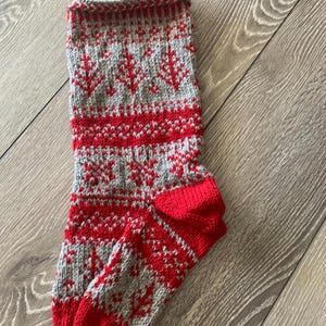May include: A hand-knitted Christmas stocking featuring a fair isle pattern in red and light grey. The design includes repeating tree and geometric motifs across the body, with a solid red heel and toe, and a small red loop at the top for hanging.