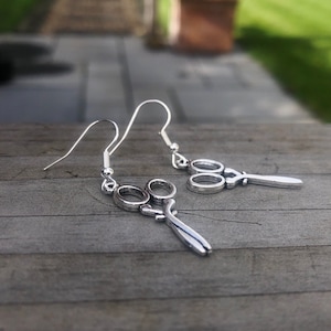 Puede incluir: Un par de pendientes colgantes de plata con un diseño de tijeras.