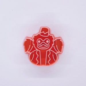Pode incluir: Um cortador de biscoitos de plástico vermelho em forma de macaco de desenho animado usando um chapéu. O macaco tem um contorno branco e um bigode branco.