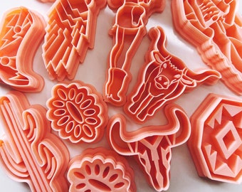 Puede incluir: Un conjunto de 11 cortadores de galletas de plástico rosa con forma de varios diseños de temática western, incluyendo un cactus, un sombrero de vaquero, una herradura, una cabeza de toro y un girasol.