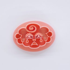 May include: A pink plastic cookie cutter shaped like a cloud with a crying face. The face has two eyes, a nose, and a mouth. The cloud has a swirl on top.