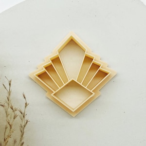 May include: A beige plastic cookie cutter shaped like a geometric sunburst design. The cutter has a square center and radiating lines that form a diamond shape.