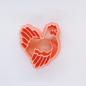 Op de afbeelding: Lichtoranje plastic koekjessteker in de vorm van een kip. De kip is in profiel afgebeeld, met gedetailleerde vleugels, een kam en een snavel. De steker is ontworpen voor het maken van koekjes of andere bakwaren.