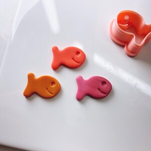 May include: Three fish-shaped cookies, one orange, one red, and one pink, on a white surface. The cookies are made with a cookie cutter in the shape of a fish.