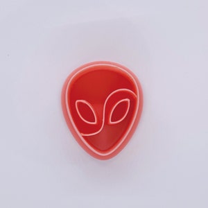 May include: A red alien head-shaped cookie cutter with a white outline. The cookie cutter is oval-shaped with two large eye shapes and a curved line for the mouth. The cutter is made of plastic and is on a white background.