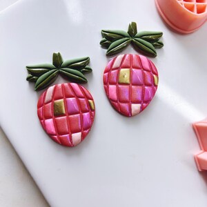 May include: Two pink and gold strawberry-shaped earrings with green leaves. The strawberries have a checkered pattern.