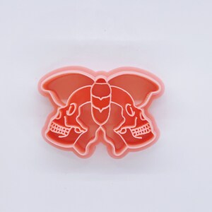 Op de afbeelding: Een roze plastic koekjesvormpje in de vorm van een vlinder met twee schedels op de vleugels.