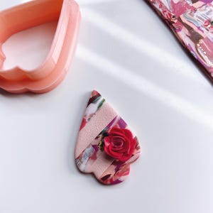 Puede incluir: Un colgante de arcilla en forma de corazón rosa y blanco con una rosa rosa y detalles dorados.