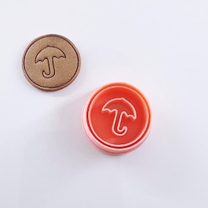 May include: A red and white plastic stamp with an umbrella design. The stamp is round and has a raised edge.