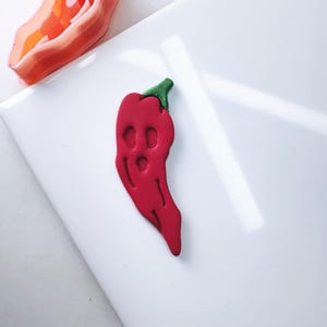 Pode incluir: Um cortador de biscoitos de pimenta vermelha com um caule verde. O cortador é feito de plástico e é projetado para cortar formas de pimenta vermelha da massa de biscoitos.