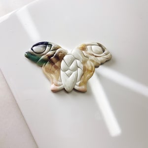 May include: A carved butterfly brooch made of white, brown, and green stone. The butterfly has intricate details and a delicate design.