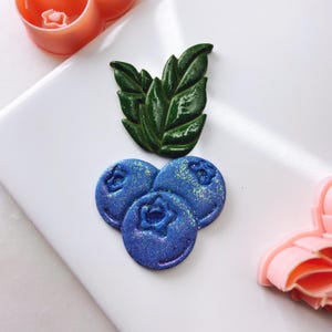 Könnte beinhalten: Drei blaue und lila glitzernde Blaubeeren mit einem grünen Blatt oben. Die Blaubeeren sind in Dreiecksform angeordnet.