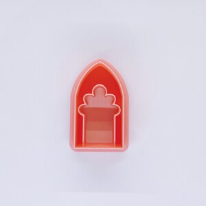 May include: A pink plastic cookie cutter shaped like an arched doorway with a decorative flower design.