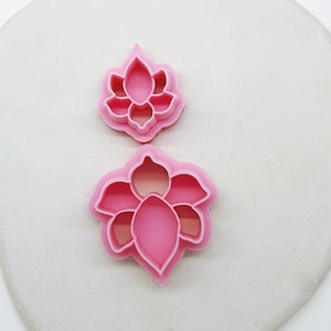 May include: Two pink plastic cookie cutters shaped like lotus flowers. The larger cutter has a more detailed design with multiple layers of petals.