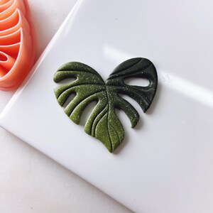 May include: A green and black glitter monstera leaf made of polymer clay.