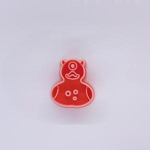 May include: A red plastic cookie cutter shaped like a cartoon monster with one eye, a mustache, and three small circles on its body.