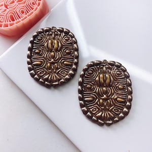 May include: Two gold-toned metal earrings with a detailed geometric design. The earrings are round and have a raised, textured surface.