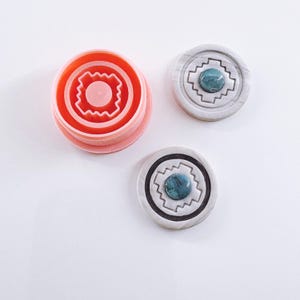 Puede incluir: Un conjunto de tres objetos redondos. Uno es un cortador de galletas de color coral con un diseño geométrico. Los otros dos son grises y blancos con un centro turquesa y un patrón geométrico. Uno tiene un borde negro.