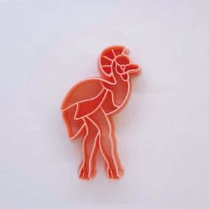 May include: A coral-colored plastic cookie cutter in the shape of a stylized ram. The cutter features a detailed design of the ram's head, body, and legs, with segmented lines defining the features. The cutter is designed for baking.